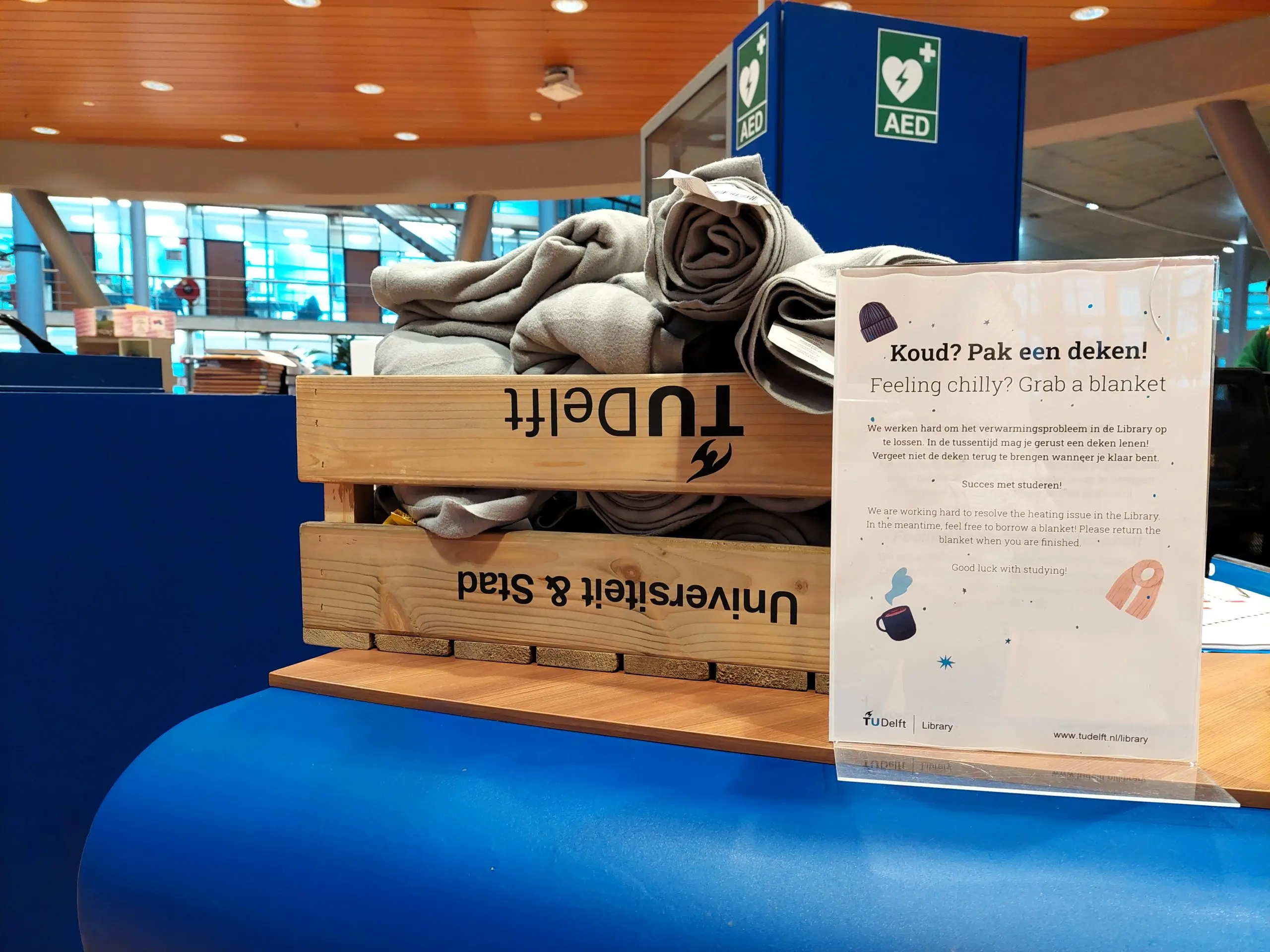 Kou_Lib_MvdV_01 Een tafel met daarop een bak vol dekentjes en de tekst 'Koud? Pak een deken'